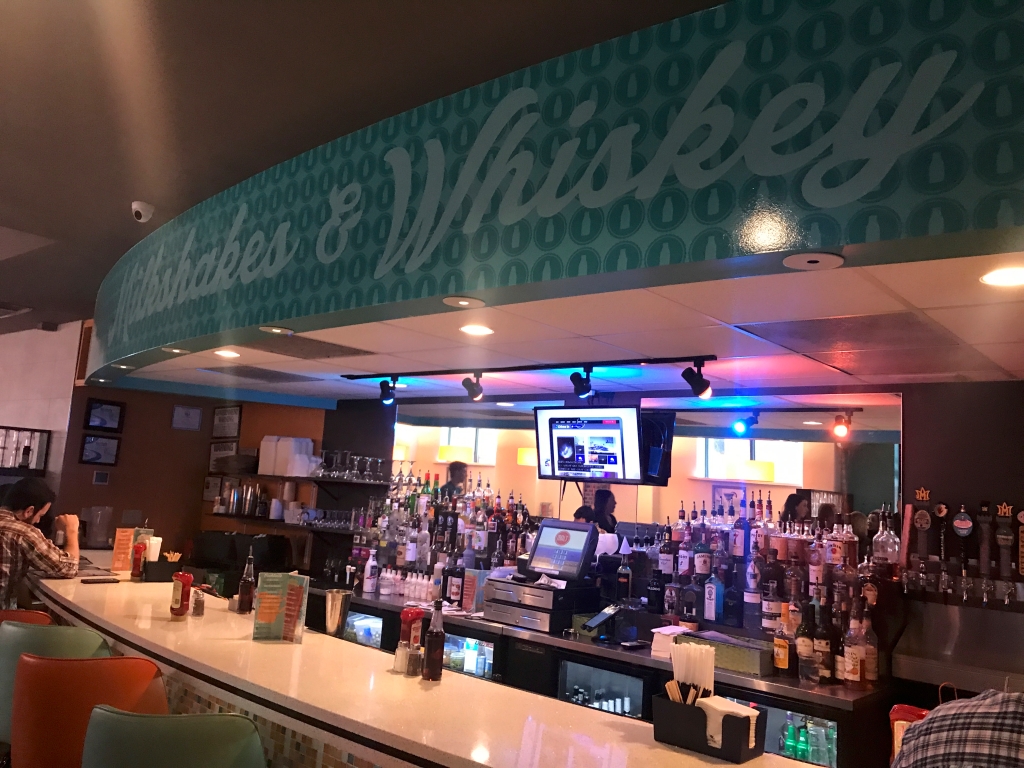 A photo of the interior inside of Modern Malt, a gastro diner in Syracuse, New York. The area above the bar is painted with words that say, "Milkshakes & Whiskey." Photo Courtesy of FoodWaterShoes