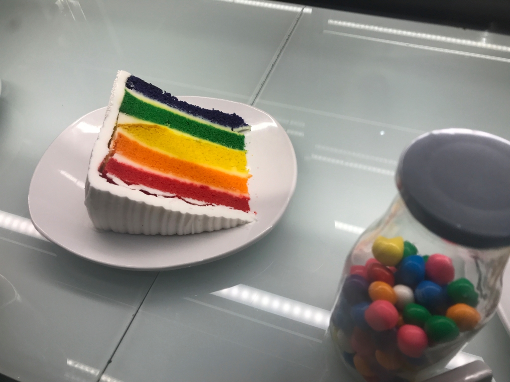 A photo of a rainbow layer cake dessert at Modern Malt in downtown Syracuse, New York. Beside the slice of cake there is a glass jar filled with colorful gumballs. Photo Courtesy of FoodWaterShoes